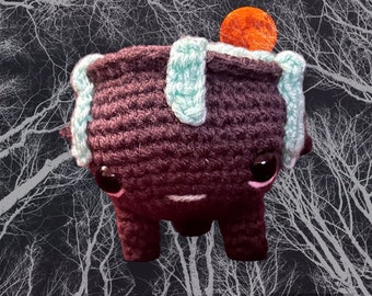 May include: A crocheted plush toy with a dark purple body, light blue accents, and large black eyes. The toy is standing in front of a dark background with white branches.