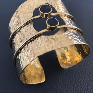 Könnte beinhalten: Ein breites, goldfarbenes Armband mit gehämmertem Muster. Zwei schwarze, kreisförmige Steine sind oben eingesetzt, verbunden durch dünne goldene Stäbe. Das Armband hat ein offenes Design und ist wahrscheinlich aus Metall.