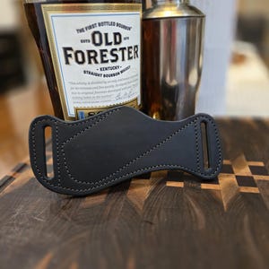Peut inclure: Un étui de bouteille de cocktail en cuir noir avec un design cousu. L'étui est à côté d'une bouteille de Old Forester Kentucky Straight Bourbon Whiskey et d'un shaker à cocktail en acier inoxydable.