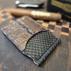 May include: A brown and black leather cigar case with crocodile and snakeskin textures. A cigar and a cigar cutter are in the background. The case is on a wooden surface.