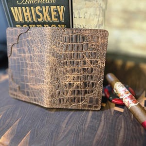 May include: A brown leather wallet with a crocodile skin texture. The wallet is open, revealing the interior. A book titled "American Whiskey" is in the background, along with a cigar and a bottle.
