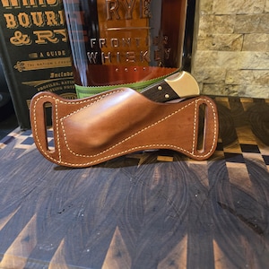 May include: A brown leather knife sheath with a knife partially inserted. The sheath has a stitched border and a curved design. A bottle of whiskey and a book are in the background.