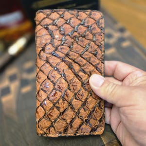 Peut inclure: Un portefeuille rectangulaire présentant un motif texturé en forme d'écailles dans des tons de brun clair et de brun foncé. La surface possède une finition brillante avec des segments distincts en losange créant un aspect organique.