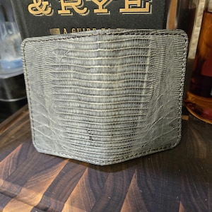 May include: A bi-fold wallet crafted from grey leather with a textured, scale-like pattern resembling lizard skin. The wallet features visible dark stitching along the edges and a rectangular shape with rounded corners, showcasing a detailed surface.
