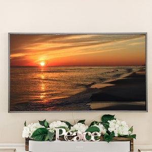 May include: A television screen showing a sunset over the ocean with a sandy beach in the foreground. The screen is mounted on a wall above a white shelf with a wooden box containing white flowers and greenery. The word "Peace" is spelled out in silver letters on the shelf.