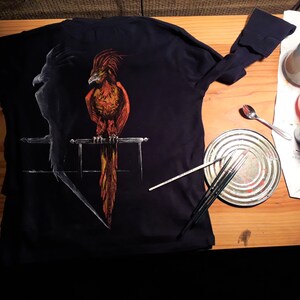 Könnte beinhalten: Ein schwarzes Langarmshirt mit einem handgemalten Design eines schwarzen Vogels und eines roten und orangefarbenen Vogels auf einer Sitzstange.