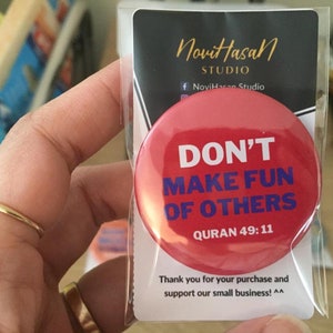 Könnte beinhalten: Roter Knopf-Abzeichen mit dem Text "DON'T MAKE FUN OF OTHERS" in weißer und blauer Schrift. Der Text "QURAN 49:11" ist in blau unter dem Haupttext gedruckt.
