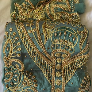 Puede incluir: Un conjunto de prendas bordadas de color verde azulado con adornos dorados. El conjunto incluye una parte superior y una bufanda transparente a juego. El bordado presenta patrones florales y geométricos, con intrincados abalorios. La tela tiene un ligero brillo.