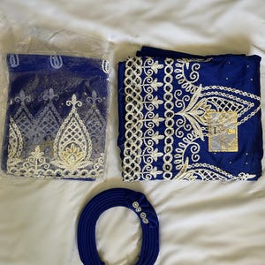 Puede incluir: Un conjunto de artículos de tela azul real, que incluye una diadema con detalles florales y dos piezas de tela bordada. Una pieza está en una bolsa de plástico. El bordado es blanco y dorado.