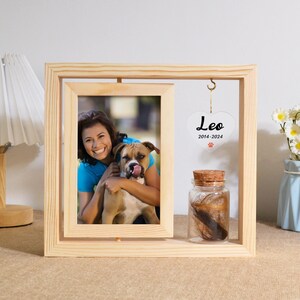 May include: Wooden picture frame with a photo of a woman and her dog. A small glass jar containing fur and a white heart with the text "Leo 2014-2024" hangs from the frame.