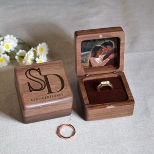 Peut inclure: Coffret à bagues en bois ouvert, révélant une bague en or avec un diamant et une photo. Le coffret est gravé des initiales "SD" et des noms "SYNTIA & DELANEY". Une bague en or rose est devant le coffret.