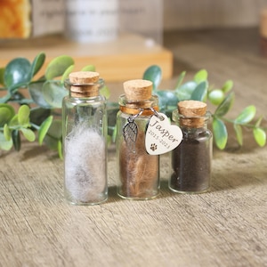 May include: Three small glass jars with cork tops, each containing a different type of pet fur. The middle jar has a silver charm attached with a paw print and the text "Jasper 2015-2023".
