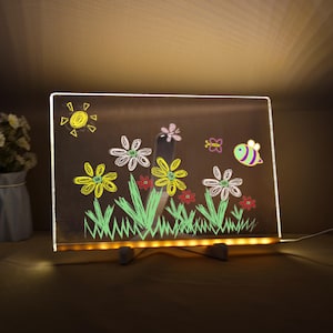 May include: An illuminated drawing board with a garden scene. The artwork includes a yellow sun, flowers, grass, a bee, and a butterfly, all drawn in chalk. The board is supported by two small white stands. The board is lit with a warm glow.