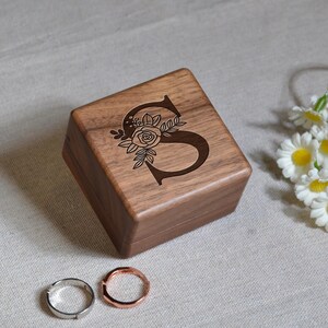 以下が含まれることがあります： 木製のリングボックスで、上面に「S」の文字が刻まれ、花の模様で飾られています。ボックスは温かみのある茶色で、銀色とローズゴールドの2つのリングの隣にあります。背景にはデイジーがあります。