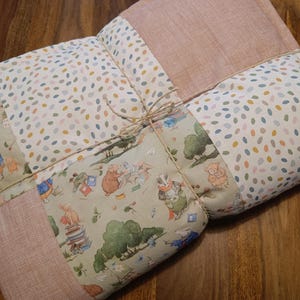 Könnte beinhalten: Eine gesteppte Decke im Patchwork-Design mit Abschnitten aus einfarbigem, altrosa Stoff, weißem Stoff mit bunten ovalen Punkten und hellgrünem Stoff, der mit Illustrationen von anthropomorphen Tieren in der Natur bedruckt ist.