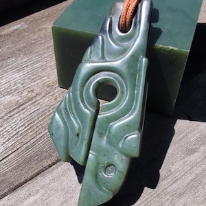May include: A dark green stone pendant featuring an intricate carved design with a central circular cutout and a vertical slit. The pendant is suspended from a brown cord that passes through a matching green stone bead at the top of the piece.