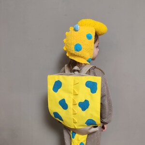 May include: A yellow crochet hat with blue spots and a dinosaur-inspired crest, paired with a matching yellow backpack featuring blue spots, a central row of triangular spikes, and a dangling tail section, worn over a brown long-sleeved garment.