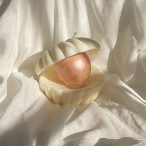 May include: A white ceramic candle shaped like a seashell with a rose gold candle inside.