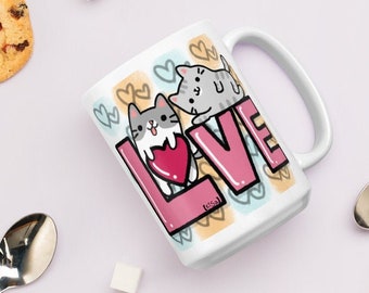May include: White ceramic mug with a pink "LOVE" graphic featuring two cartoon cats. The cats are holding a pink heart and surrounded by hearts and other doodles.