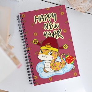May include: A red spiral notebook featuring a cartoon snake wearing a red and gold hat. The notebook has a red background with gold coins and the words "Happy New Year" in white.