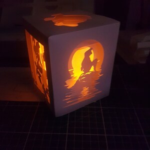 Könnte beinhalten: Eine weiße Papierlaterne mit einer Silhouette einer Meerjungfrau, die auf einem Felsen sitzt, mit einem Vollmond hinter ihr. Die Laterne wird von innen beleuchtet und strahlt ein warmes Licht aus.