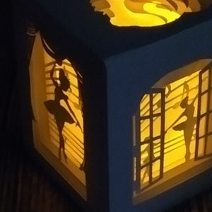 May include: A blue, cube-shaped paper art piece with cut-out silhouettes of ballerinas and a swan. The interior is illuminated with a warm, yellow light, creating a layered effect. The ballerina cutouts are positioned to look like they are dancing in front of a window.