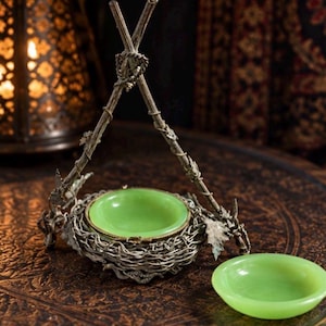 May include: A decorative metal stand featuring a twig-like frame with a central nest-shaped holder containing a smooth, round, light green bowl. A matching light green bowl sits separately in front of the structure, which is crafted from silver-toned metal.