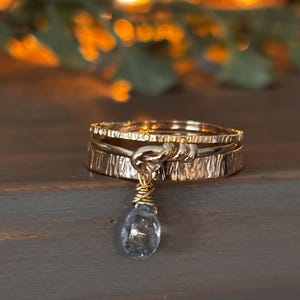 Puede incluir: Un anillo de oro con un delicado colgante de piedra azul en forma de lágrima. El anillo está hecho de dos bandas con un acabado texturizado.