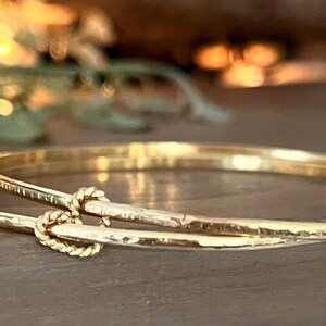 May include: Two gold bangles with a knotted design. The bangles are stacked on top of each other and have a hammered texture.