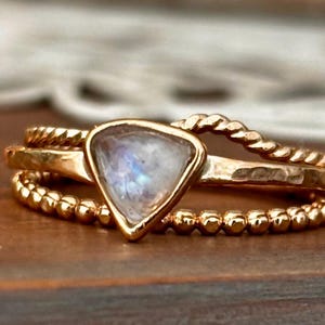 May include: A trio of gold rings. The top ring has a twisted rope design, the middle ring has a hammered texture, and the bottom ring is beaded. The center ring features a triangular moonstone.