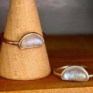 May include: Two rings featuring semi-circular moonstone cabochons set in metal. One ring has a gold-toned band and setting, while the other has a silver-toned band and setting. The stones display a translucent, iridescent quality with blue hues.