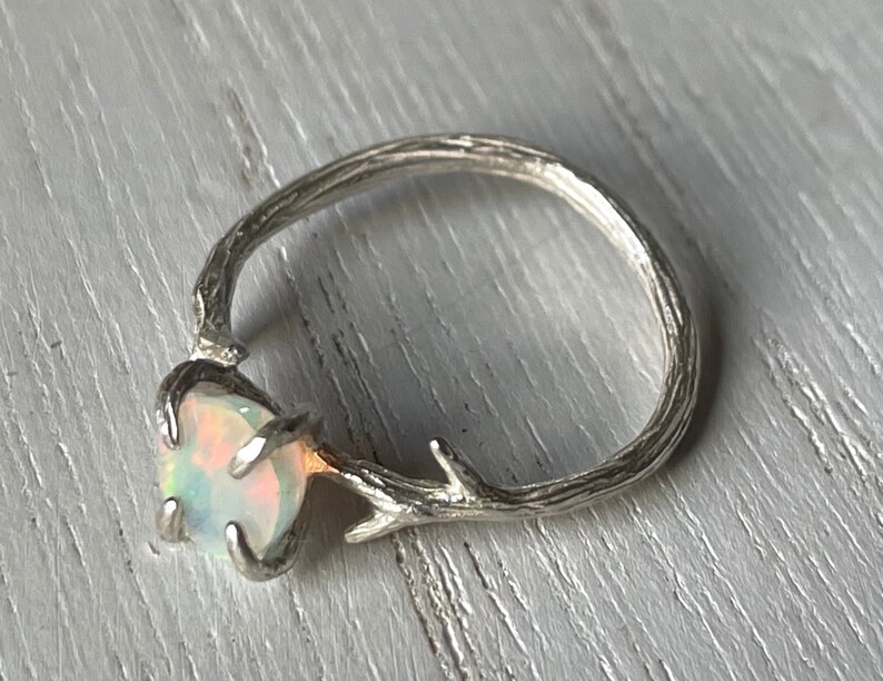 May include: A silver ring with a branch design and a white opal gemstone. The ring is made to look like a twig with a small opal set in the center.