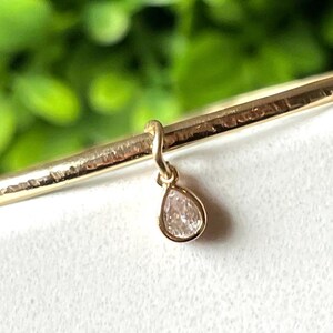 Peut inclure: Gros plan sur un bracelet doré avec un pendentif en forme de poire, de couleur claire. Le bracelet a une texture martelée. L'arrière-plan est flou, avec du feuillage vert et une surface blanche.