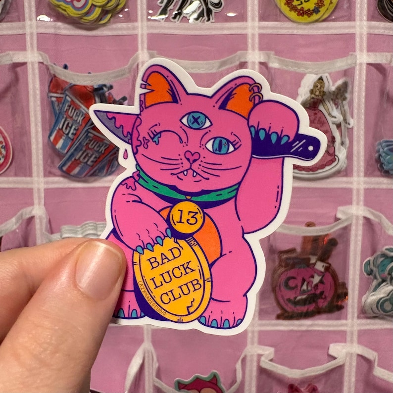Puede incluir: Una pegatina rosa con un gato de dibujos animados con tres ojos, gui&ntilde;ando un ojo y sosteniendo un peque&ntilde;o objeto negro. El gato tiene la cabeza naranja y un collar verde. La pegatina tiene una etiqueta dorada que dice "BAD LUCK CLUB" y el n&uacute;mero "13".