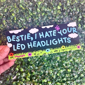 Puede incluir: Una pegatina rectangular con fondo azul oscuro, nubes blancas y el texto en azul claro 'BESTIE, I HATE YOUR LED HEADLIGHTS'. El borde inferior muestra hierba verde con flores coloridas, un gusano rosa y el texto 'LAZY MADE STUDIO'.
