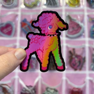 May include: A colorful sticker of a lamb with a gradient of pink, orange, green, and purple. The lamb has a spiked collar and a heart tattoo with the word "ME". The sticker is held by a hand.
