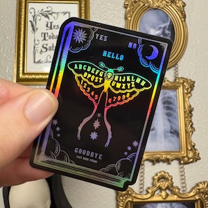 Puede incluir: Una pegatina holográfica negra con un diseño de polilla, las palabras "HELLO" y "GOODBYE", el alfabeto, números y las palabras "YES" y "NO". La pegatina es de Lazy Made Studio.