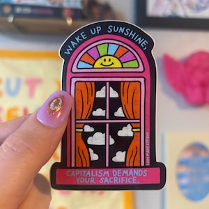 May include: A die-cut sticker featuring a pink window frame with orange curtains, a rainbow arch, and a yellow smiley face sun. The text reads WAKE UP SUNSHINE, at the top and CAPITALISM DEMANDS YOUR SACRIFICE. at the bottom against a black background.