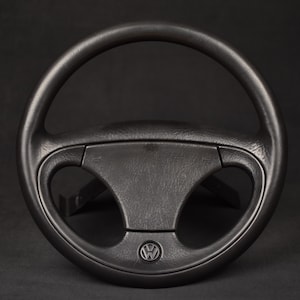 Könnte beinhalten: Ein schwarzes Volkswagen-Lenkrad mit einem kreisförmigen Kranz und einem zentralen Pralltopf, auf dem unten das ikonische VW-Logo eingeprägt ist. Das Lenkrad hat eine strukturierte Oberfläche und ein Drei-Speichen-Design mit zwei großen Öffnungen.