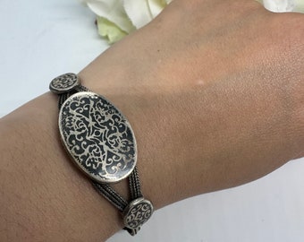 May include: A silver bracelet with a large oval charm featuring a black and silver filigree design. The bracelet has two smaller round charms with a similar design.