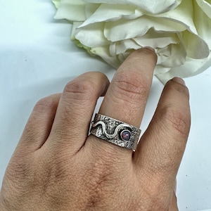 Könnte beinhalten: Ein silberner Ring mit einem strukturierten Band und einem kleinen rosafarbenen Opal-Stein. Das Band hat ein wellenförmiges Design.