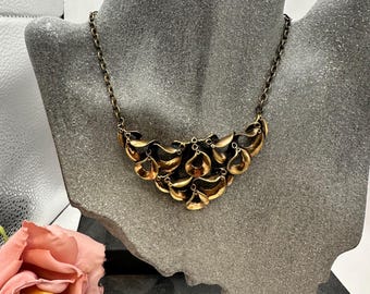 Peut inclure: Un collier doré au design unique, présenté sur un buste gris. Le collier présente une série de formes stylisées en forme de pétales. La chaîne est en métal foncé et délicat. Une rose rose est visible dans le coin inférieur gauche.