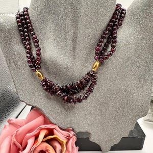 Op de afbeelding: Een ketting met meerdere strengen, weergegeven op een grijze buste. De ketting heeft drie strengen donkerrode kralen en een centraal gedeelte van ruw geslepen, donkerrode stenen. Goudkleurige sluitingen verbinden de strengen. Een roze roos staat op de voorgrond.
