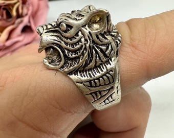 May include: A silver ring with a detailed tiger head design. The tiger's face is sculpted with intricate lines, showing its teeth and fur. The ring is worn on a finger, highlighting the detailed craftsmanship of the jewellery.