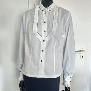 Puede incluir: Camisa blanca de manga larga con botones, cuello alto y ribete de encaje. La camisa tiene un cuello y puños con volantes.