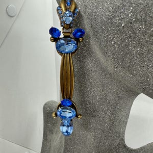 Puede incluir: Un pendiente largo y ornamentado con una base de metal dorado y múltiples detalles de cristal azul. El pendiente presenta una parte superior en forma de corona, cristales azules ovalados y redondos, y un diseño vertical acanalado. El estilo general es vintage y elegante.