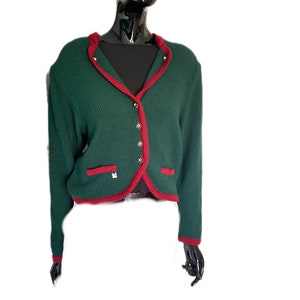 Könnte beinhalten: Ein grüner Strickcardigan mit rotem Besatz und goldenen Knöpfen. Der Cardigan hat einen V-Ausschnitt und lange Ärmel.