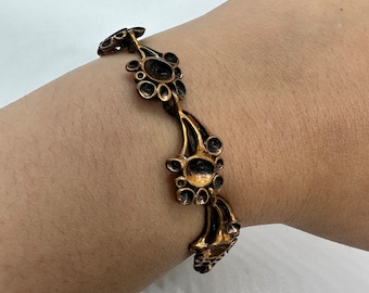 May include: A gold-toned bracelet with a floral design. The bracelet is made of metal and has a series of connected circles and swirls.