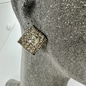 Puede incluir: Un par de pendientes cuadrados de tono plateado con diamantes de imitación transparentes. Los pendientes tienen un gran diamante de imitación transparente en el centro rodeado de diamantes de imitación más pequeños.