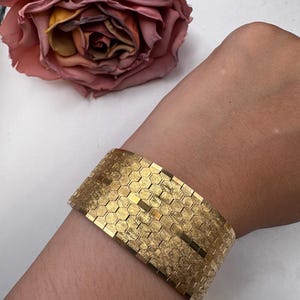 Könnte beinhalten: Ein breites Goldarmband mit einem sich wiederholenden Waben- oder Sechseckmuster und einer strukturierten Oberfläche. Die Metallglieder sind in mehreren horizontalen Reihen angeordnet und bilden ein flexibles Band am Handgelenk.
